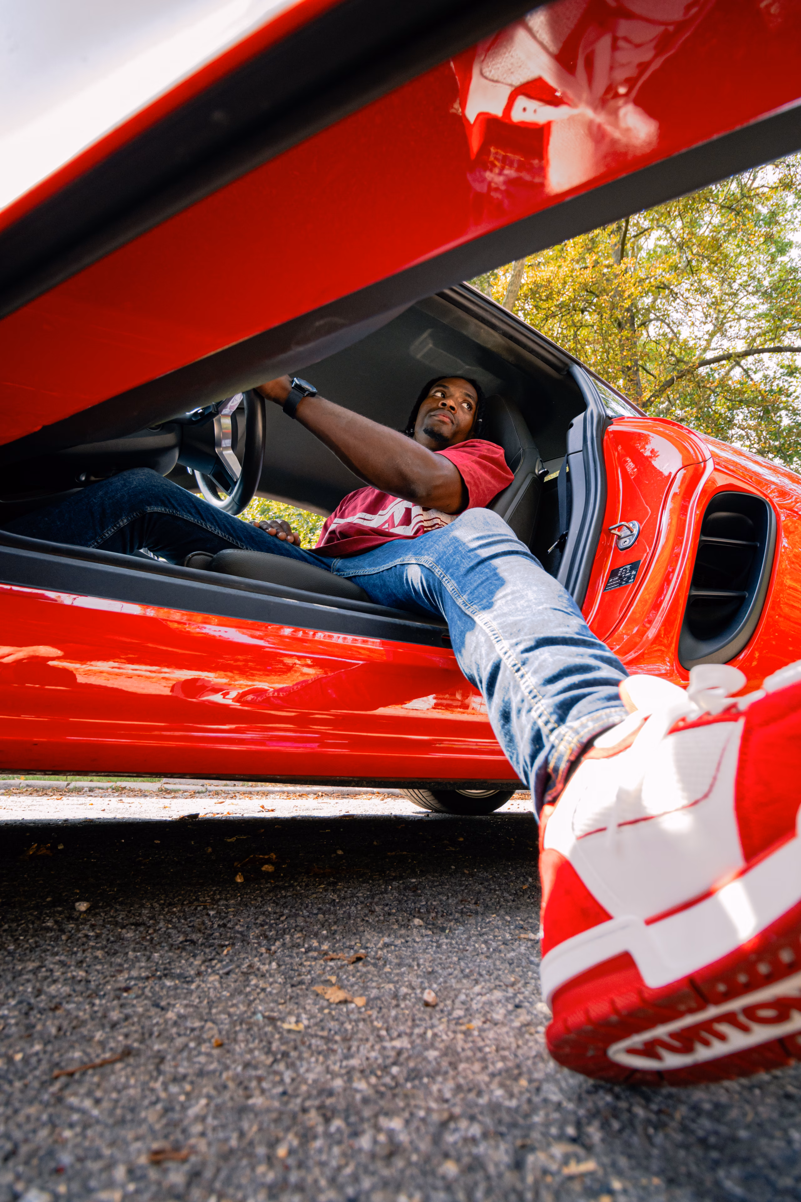 Homme installé au volant d'une voiture de sport rouge, portière relevée et jambe tendue vers l'objectif