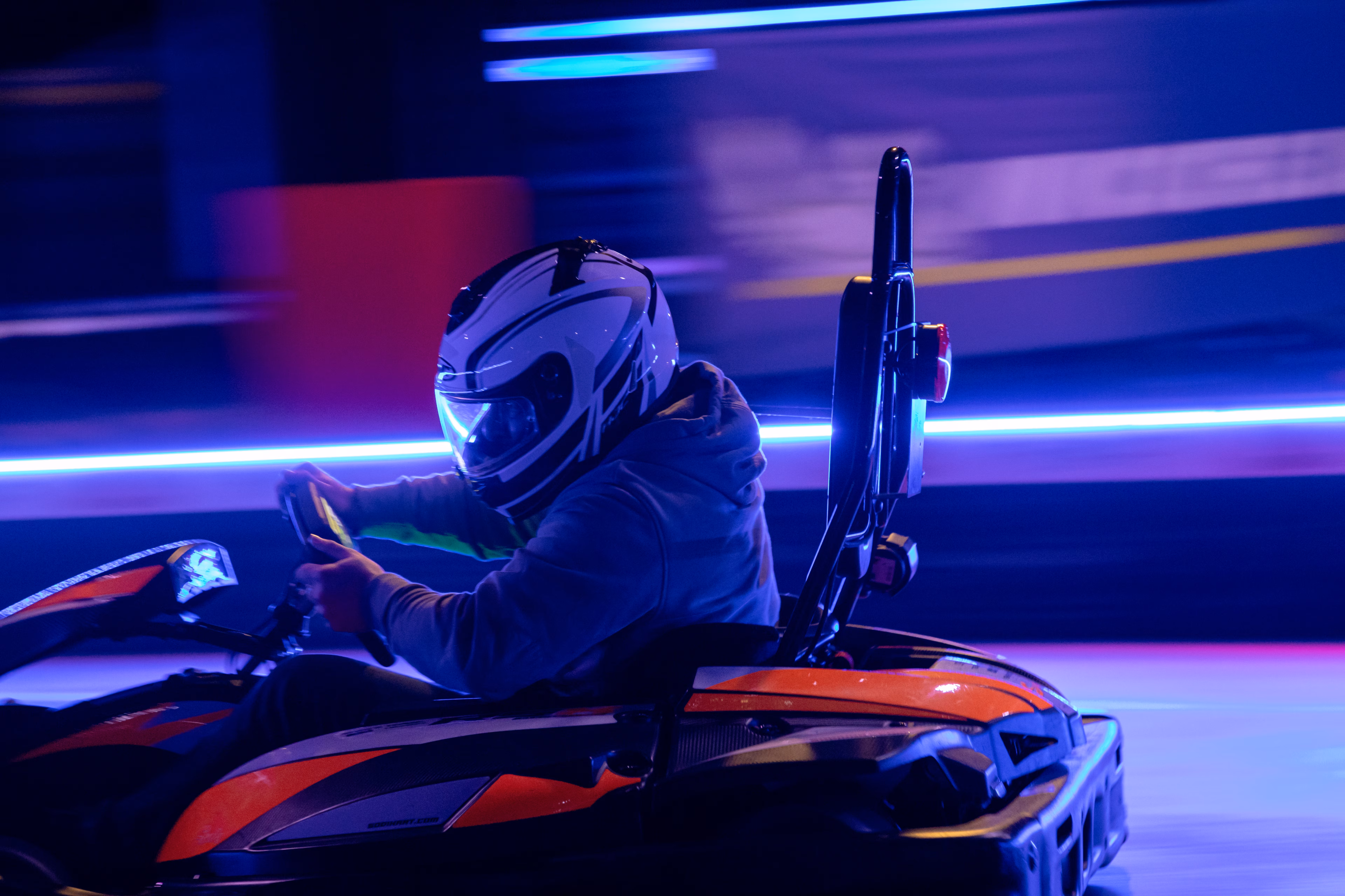 Pilote de kart en casque blanc en plein virage, néons bleus derrière lui