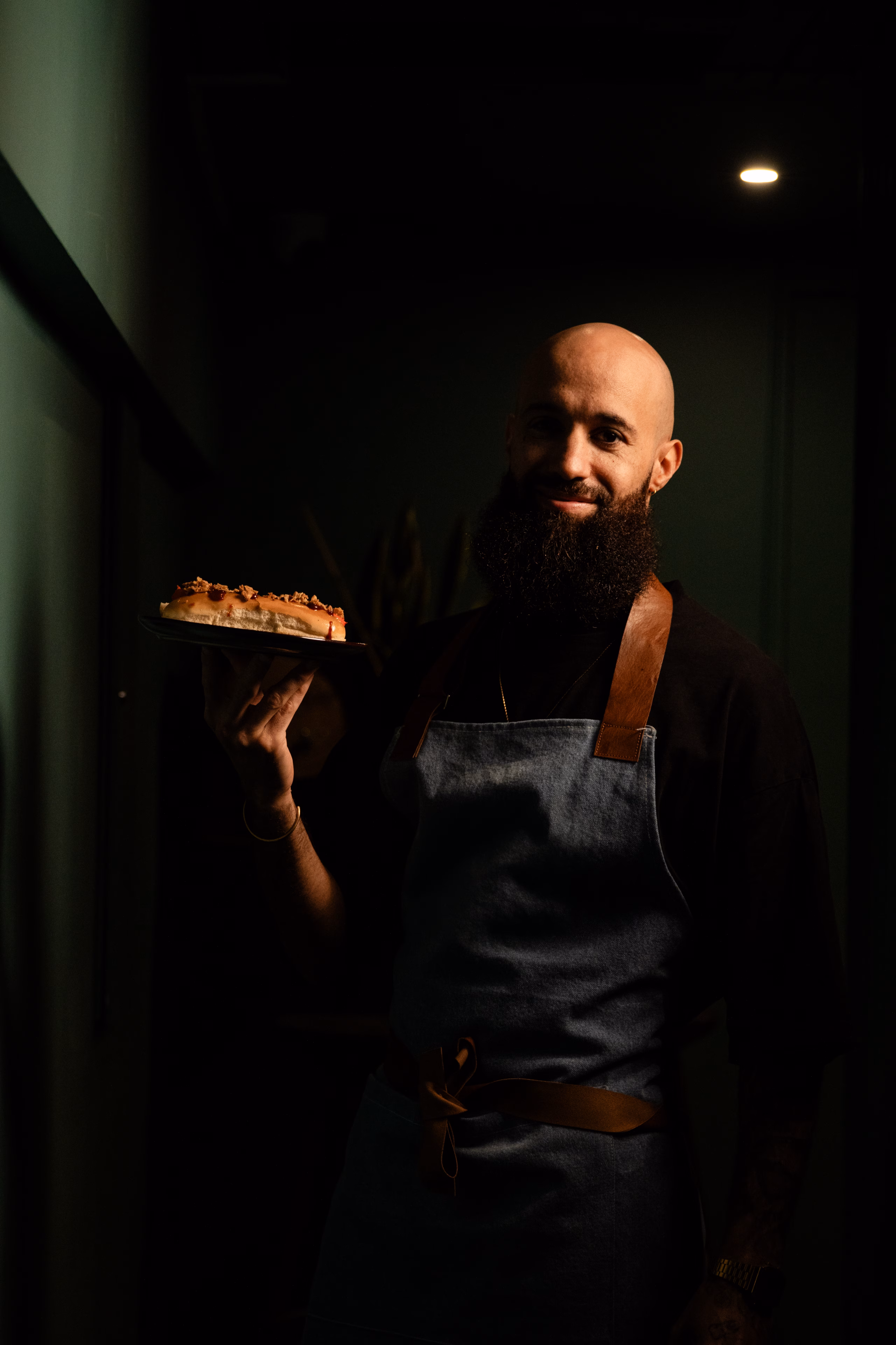 Serveur barbu en tablier de jean présentant une assiette dans la pénombre du restaurant
